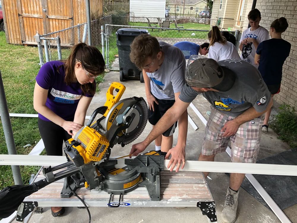 North Shore high schoolers in safety glasses measuring and cutting siding on a miter saw during their weeklong working vacation.